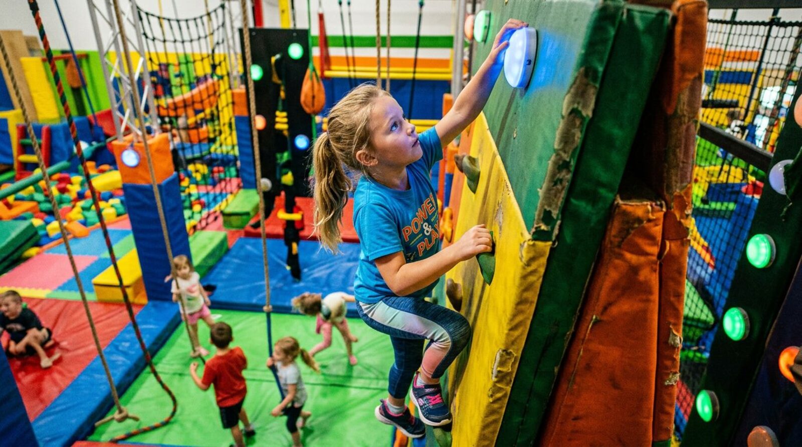 A girl climbing an indoor obstacle wall to hit a glowing pod at the top